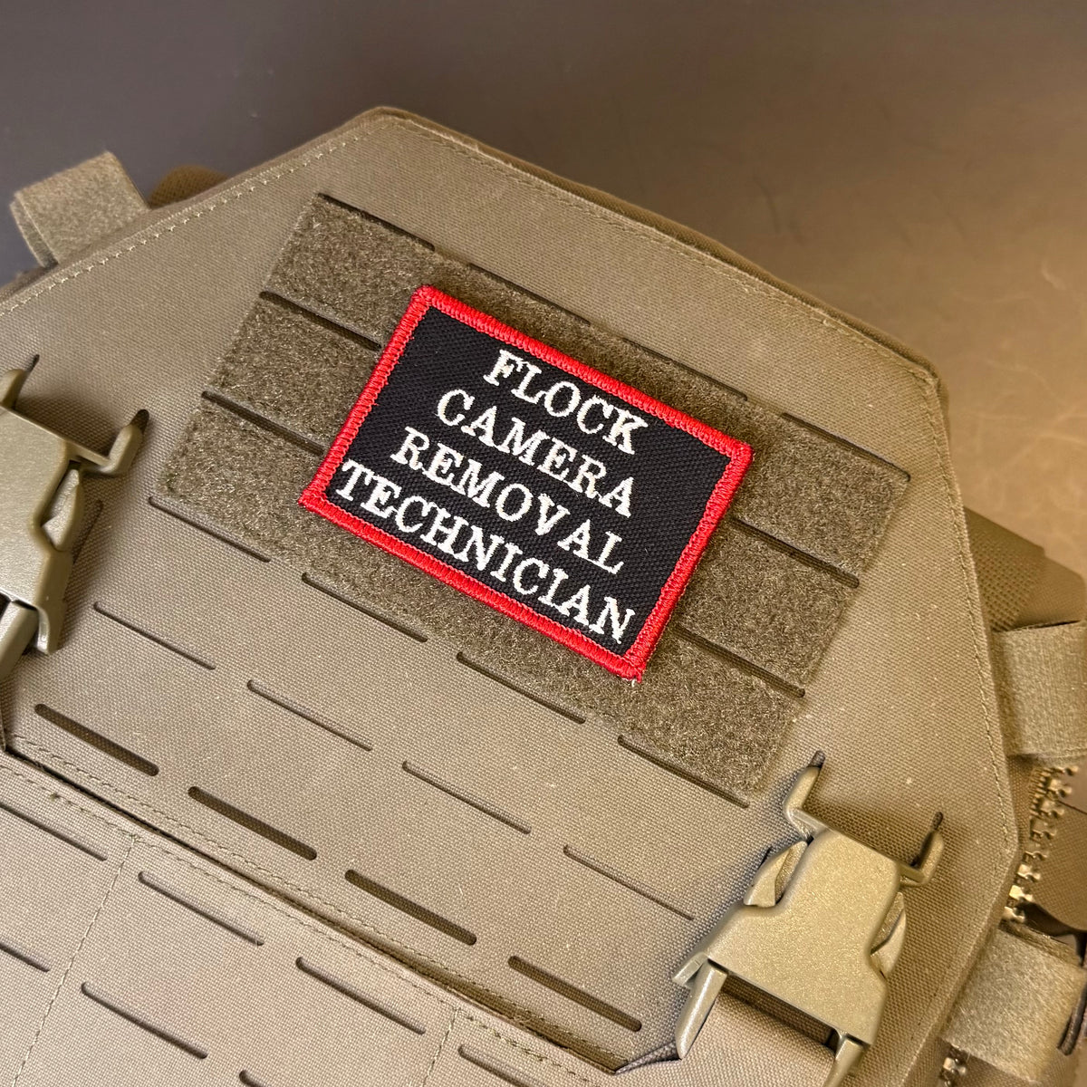 Flock Camera Removal Technician embroidered morale patch: with a black background, red border, and white text reading 'FLOCK CAMERA REMOVAL TECHNICIAN,' attached to a tan tactical vest or bag with hook-and-loop.
