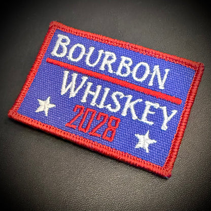 A rectangular embroidered patch with a blue field, red border, white stars, and white and red text reading “BOURBON WHISKEY 2028,” photographed on a dark textured surface.