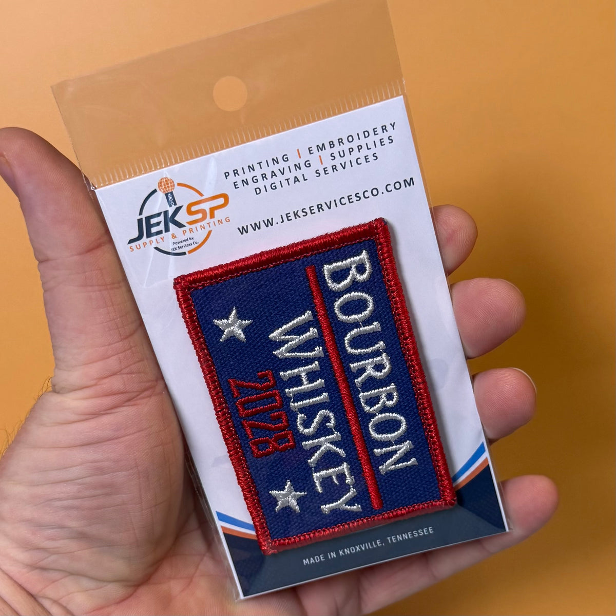 Bourbon & Whiskey for President 2028 embroidered morale patch: hand holds a packaged embroidered patch that reads "Bourbon Whiskey 1798" in white and red on a dark blue background. The patch is attached to a white backing card.