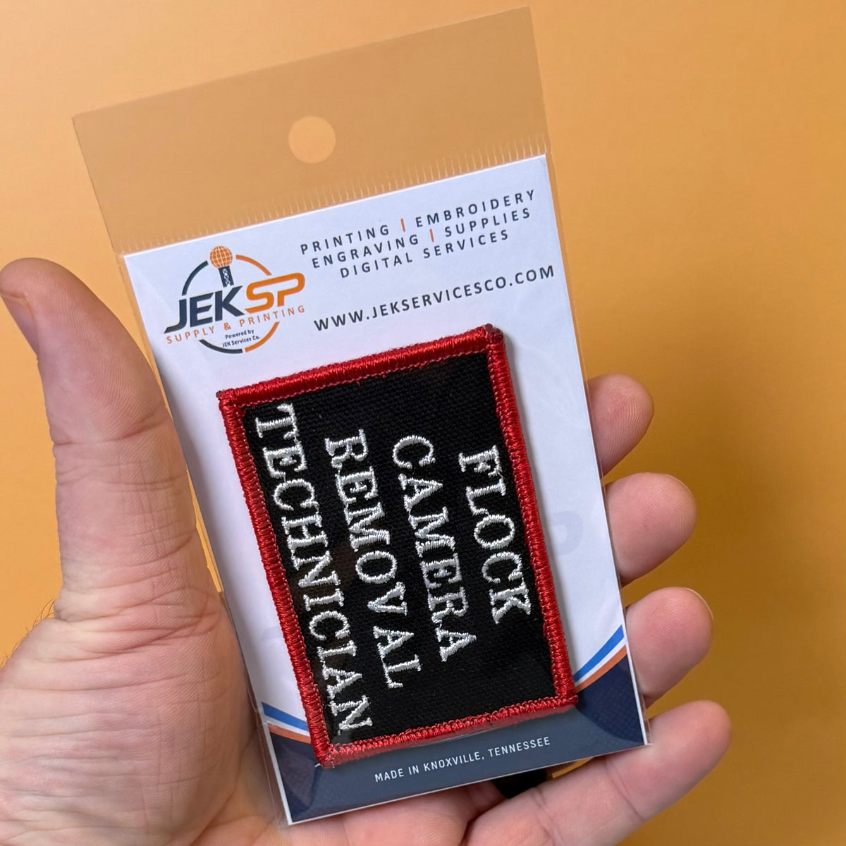 Flock Camera Removal Technician embroidered morale patch: hand holding a packaged embroidered patch that reads 'FLOCK CANVAS REMOVAL TECHNICIAN' in white text on a black background with a red border, against a solid orange.