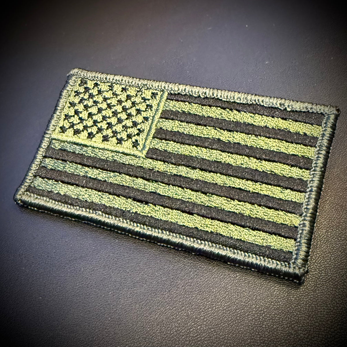 An embroidered American flag patch in olive drab and black, shown on a dark textured surface.