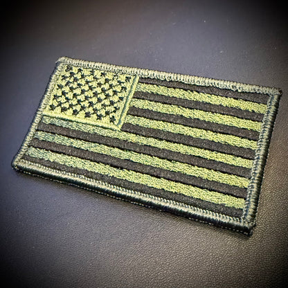 An embroidered American flag patch in olive drab and black, shown on a dark textured surface.