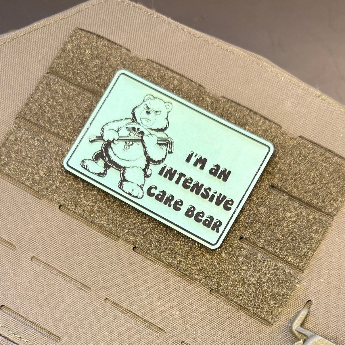 Intensive Care Bear Funny Nurse Medic laser-engraved synthetic morale patch: a rectangular tactical patch featuring a cartoon bear holding a thermometer with the text 'I'M AN INTENSIVE CARE BEAR', attached to tan webbing on a beige.