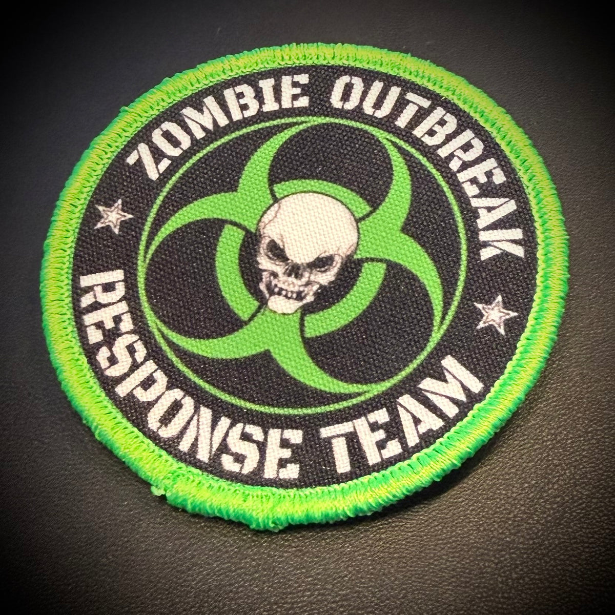 A circular embroidered patch with a bright green border, black center, white text reading 'ZOMBIE OUTBREAK RESPONSE TEAM,' a green biohazard symbol, and a white skull in the middle, shown on a dark textured background.