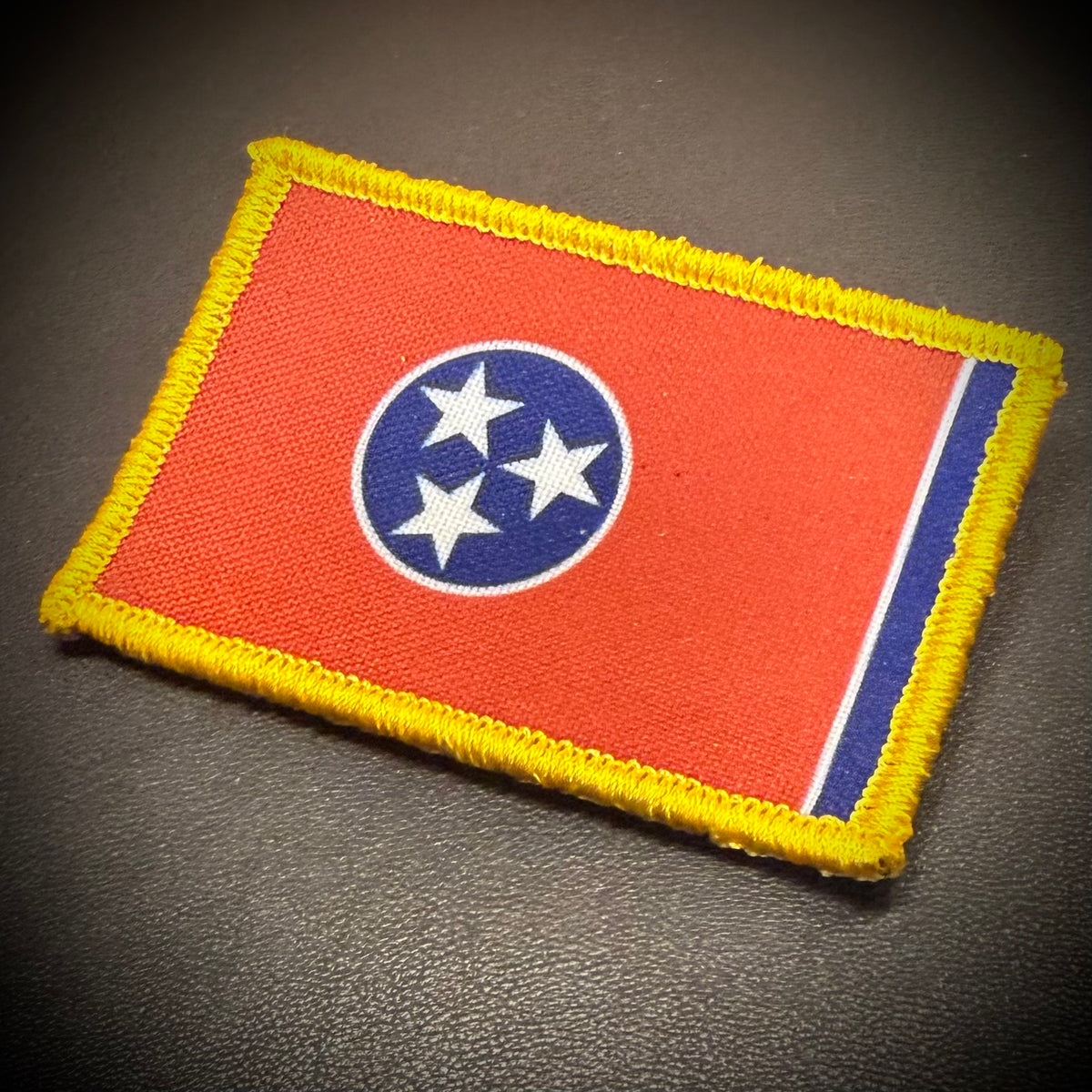 An embroidered rectangular patch featuring the Tennessee state flag design, with a red field, blue circle with three white stars, and a yellow border, photographed on a dark textured surface.