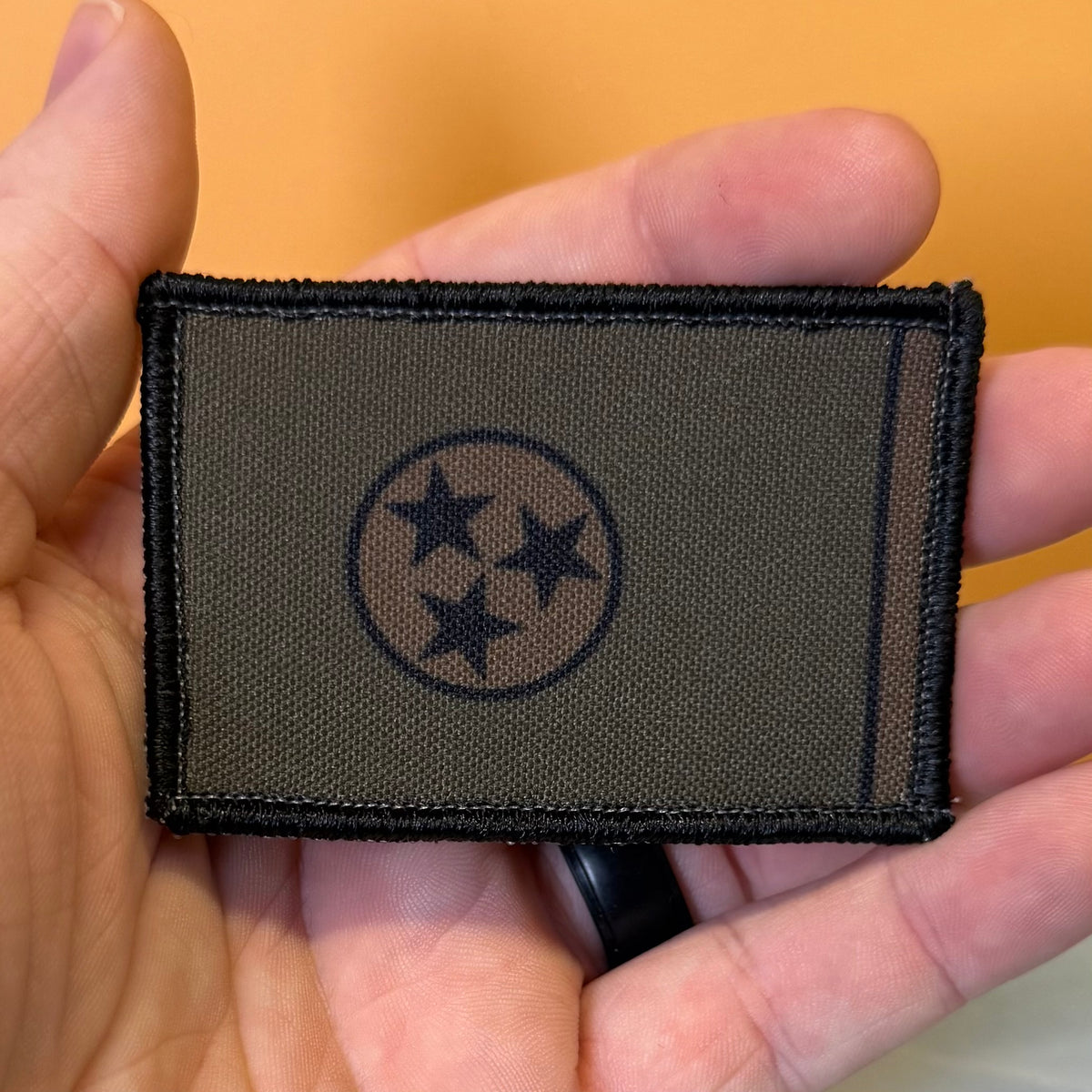 Tennessee State Flag printed canvas morale patch featuring the Tennessee state flag design in olive drab and black, held in a person's hand against a solid orange background.