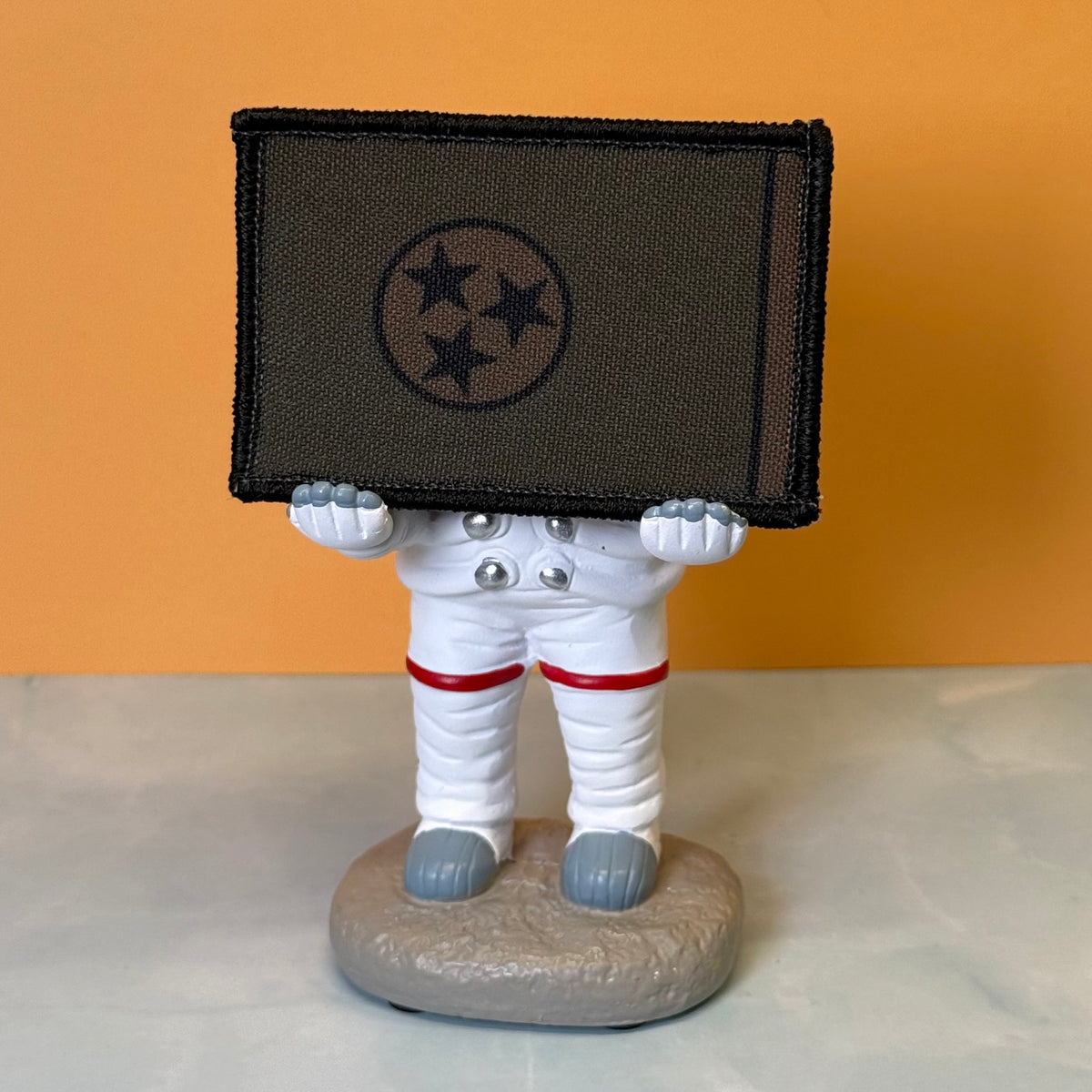 Tennessee State Flag printed canvas morale patch, a small astronaut figurine holding a dark rectangular patch with three black stars in a circle, set against an orange background and light gray surface.