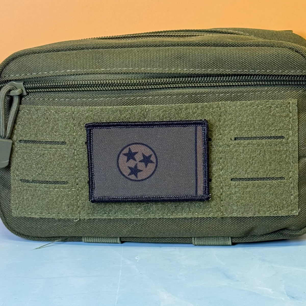 Tennessee State Flag printed canvas morale patch, an olive green tactical pouch with a front zipper and a dark gray rectangular patch featuring a black three-star circle emblem, set against a split orange and light blue.