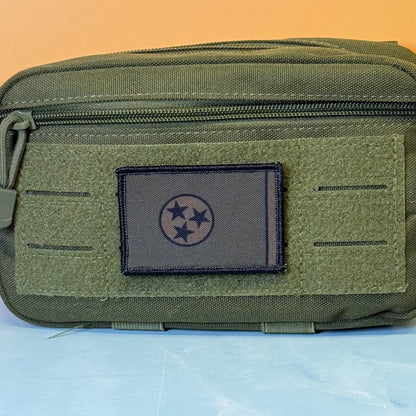 Tennessee State Flag printed canvas morale patch, an olive green tactical pouch with a front zipper and a dark gray rectangular patch featuring a black three-star circle emblem, set against a split orange and light blue.