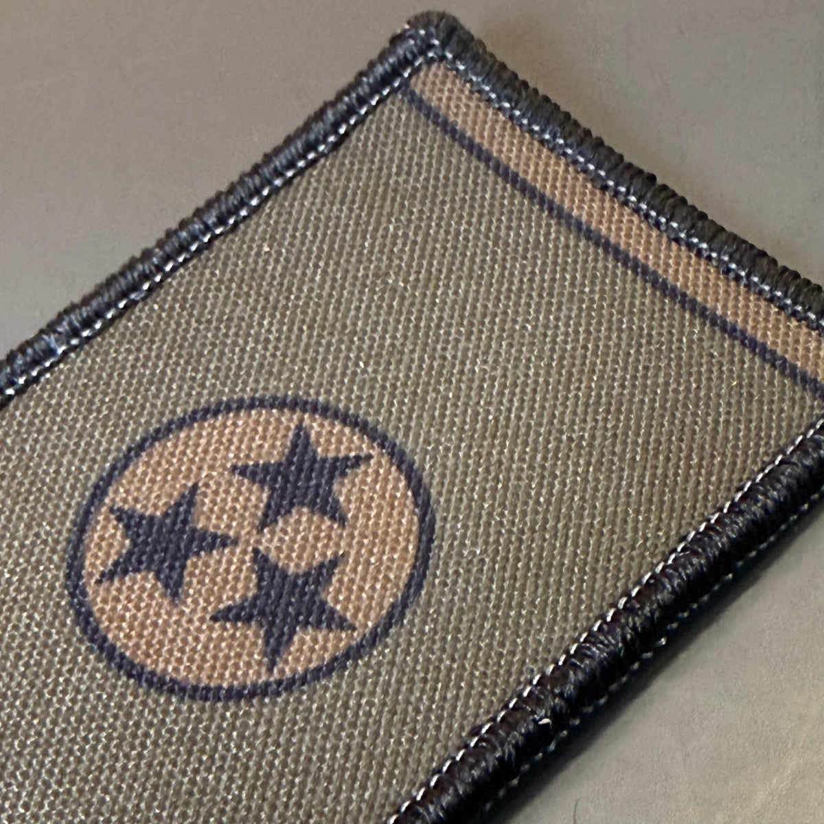 Tennessee State Flag printed canvas morale patch, close-up of a rectangular woven patch featuring a tan circle with three dark blue stars, bordered by dark stitching and a tan strip near the top edge.