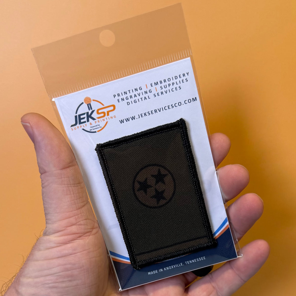 Tennessee State Flag printed canvas morale patch, a hand holds a packaged black rectangular printed patch featuring a circular Tennessee state flag design. The patch is mounted on a white backing card with the JEKSP logo and.