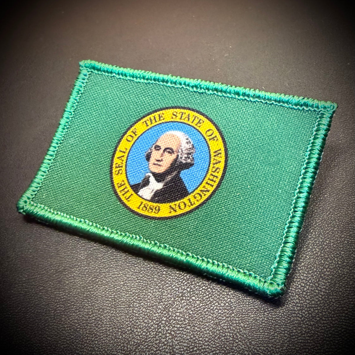 A green embroidered patch featuring the seal of the State of Washington, displayed on a dark textured surface.