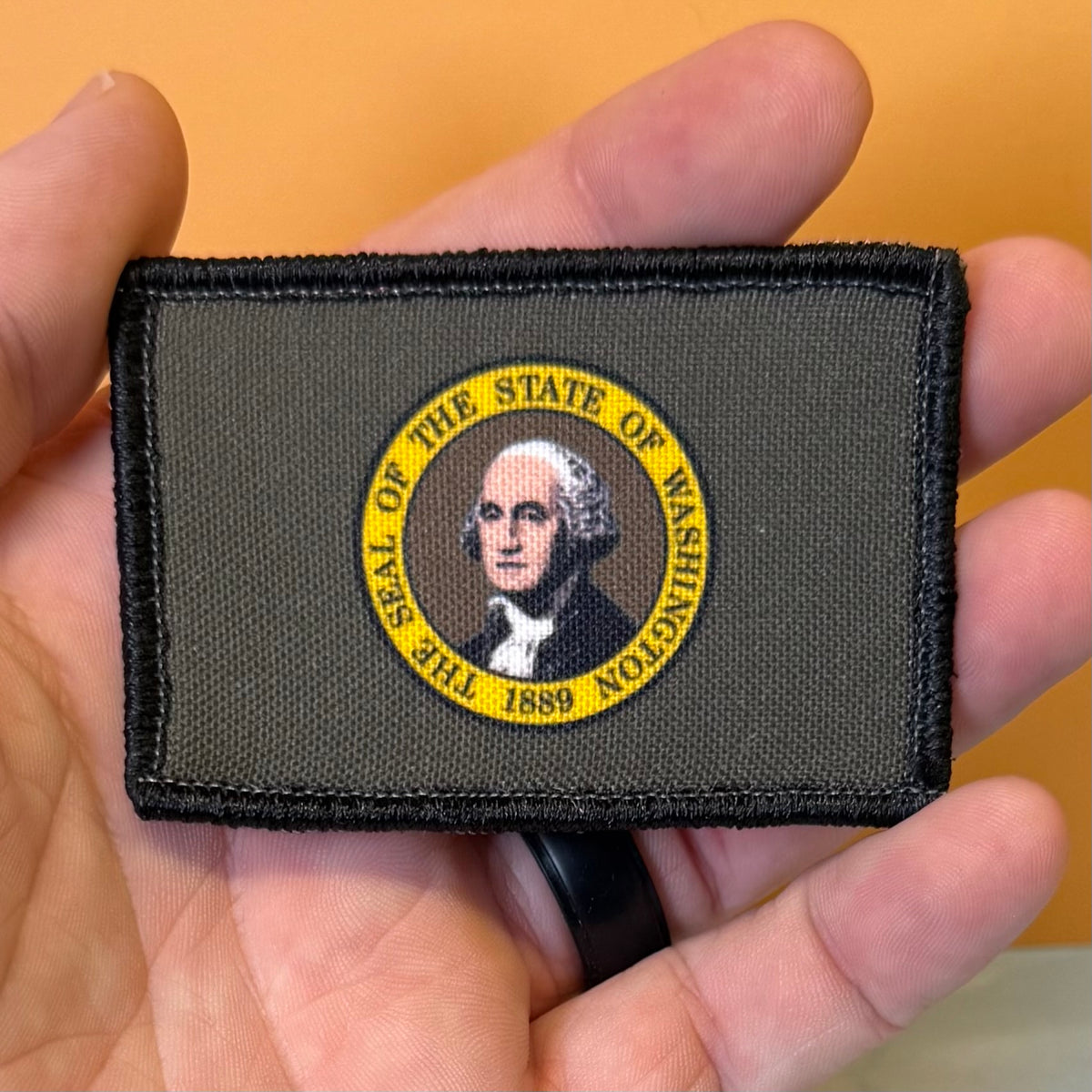 Washington State Flag printed canvas morale patch featuring the Seal of the State of Washington, held in a person's hand against a solid orange background.
