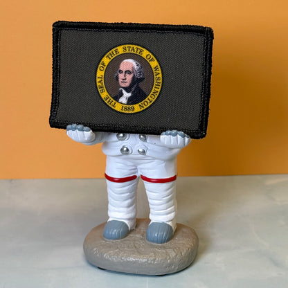 Washington State Flag printed canvas morale patch, a small astronaut figurine holding a black rectangular patch featuring the Seal of the State of Washington with a portrait of George Washington and yellow text. The figurine.