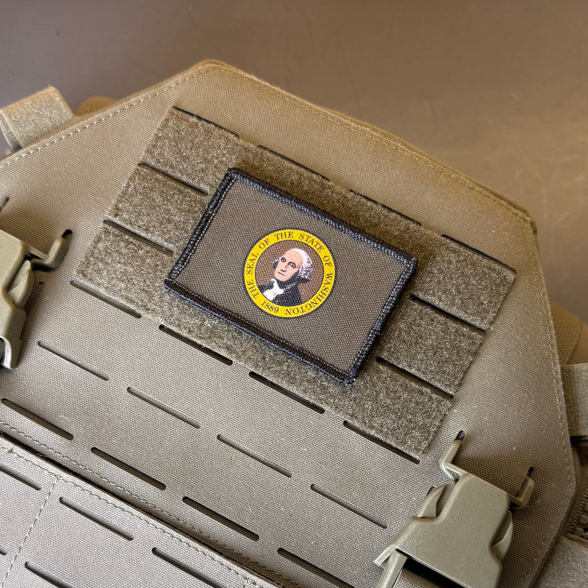Washington State Flag printed canvas morale patch featuring a portrait of George Washington with a yellow circular border and the design lettering and 'IN NO TREATY', mounted on a tan MOLLE webbing strap of a tactical vest or bag.