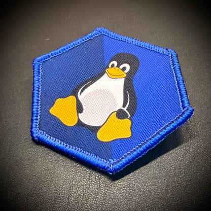 A hexagonal embroidered patch featuring the Tuxedo penguin logo on a blue background with a blue stitched border, placed on a dark textured surface.