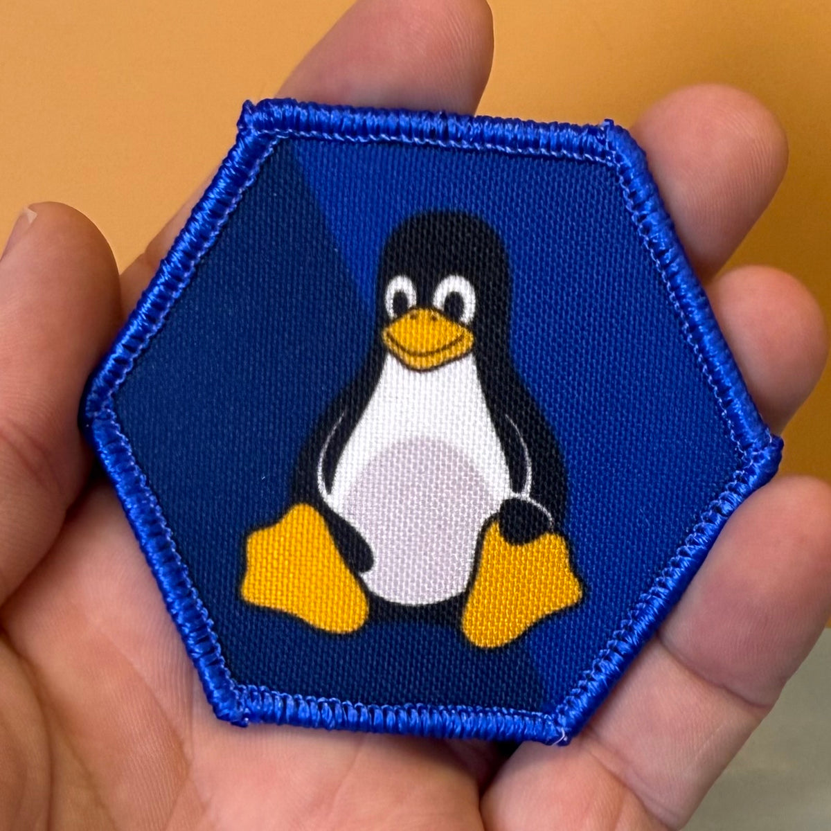 Linux Tux Penguin printed canvas morale patch featuring the Tuxedo penguin logo on a blue background, held in a person's hand against a warm orange background.