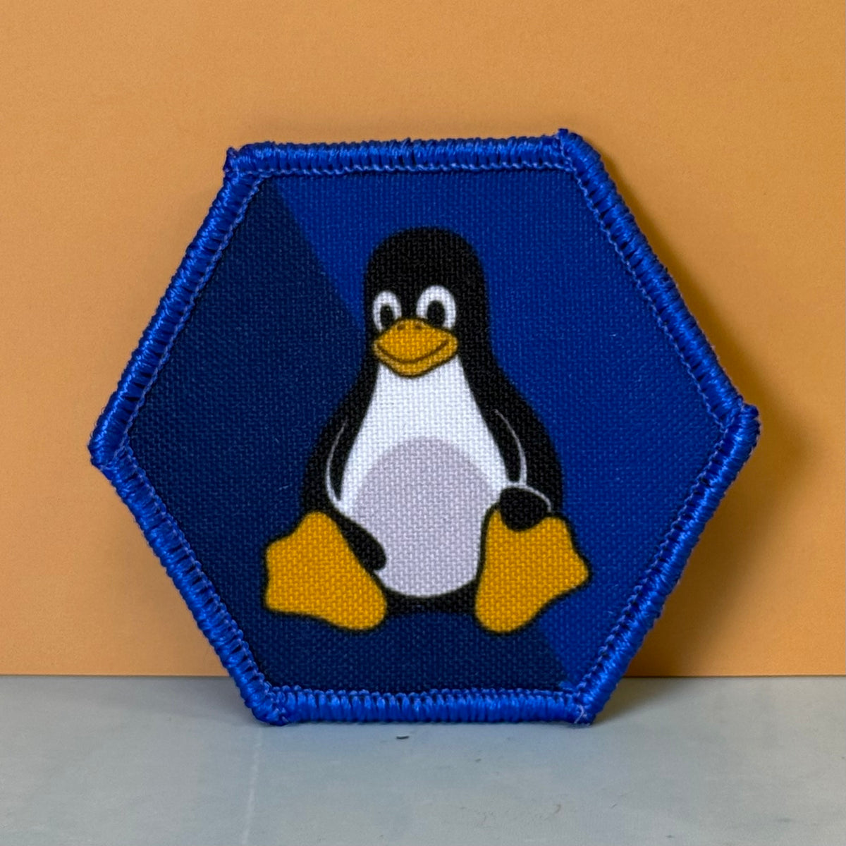 Linux Tux Penguin printed canvas morale patch featuring the Tuxedo penguin on a blue background with a blue stitched border, set against a warm orange background.