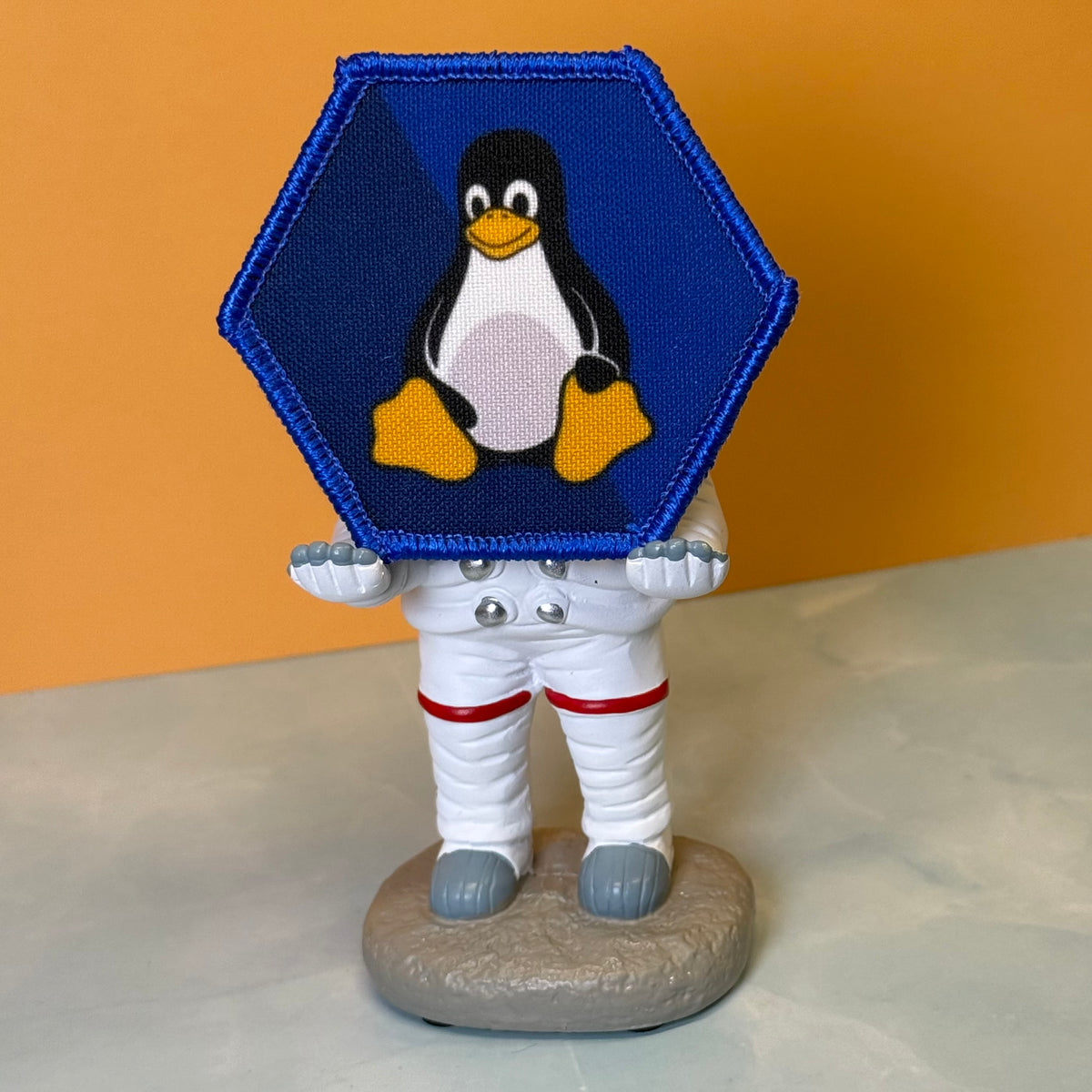 Linux Tux Penguin printed canvas morale patch, a small astronaut figurine standing on a round base, holding a large hexagonal blue patch with a penguin logo. The astronaut is dressed in a white spacesuit with red stripes and.