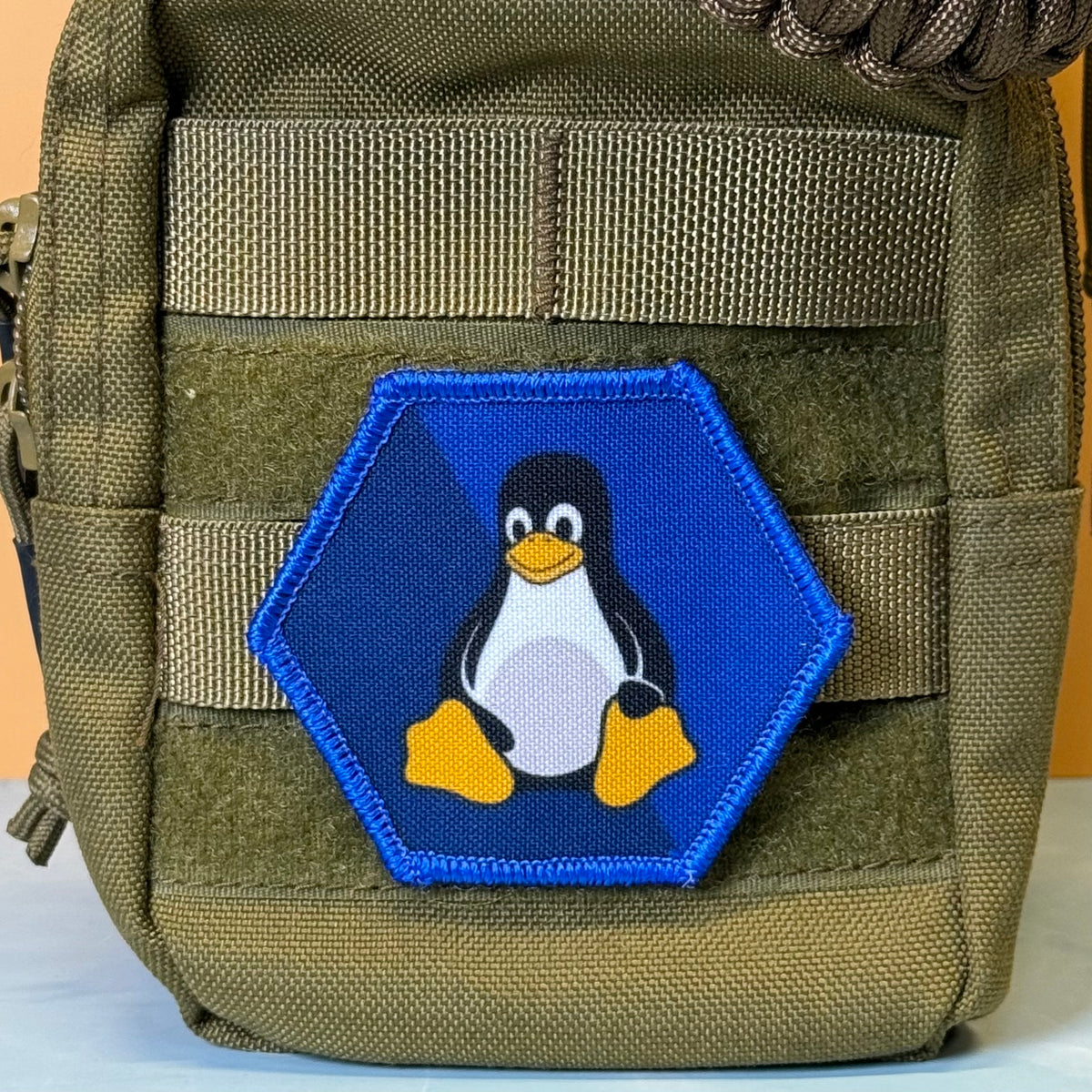 Linux Tux Penguin printed canvas morale patch, a close-up of an olive green tactical pouch featuring a hexagonal printed patch with the Tuxedo Linux penguin logo on a blue background.