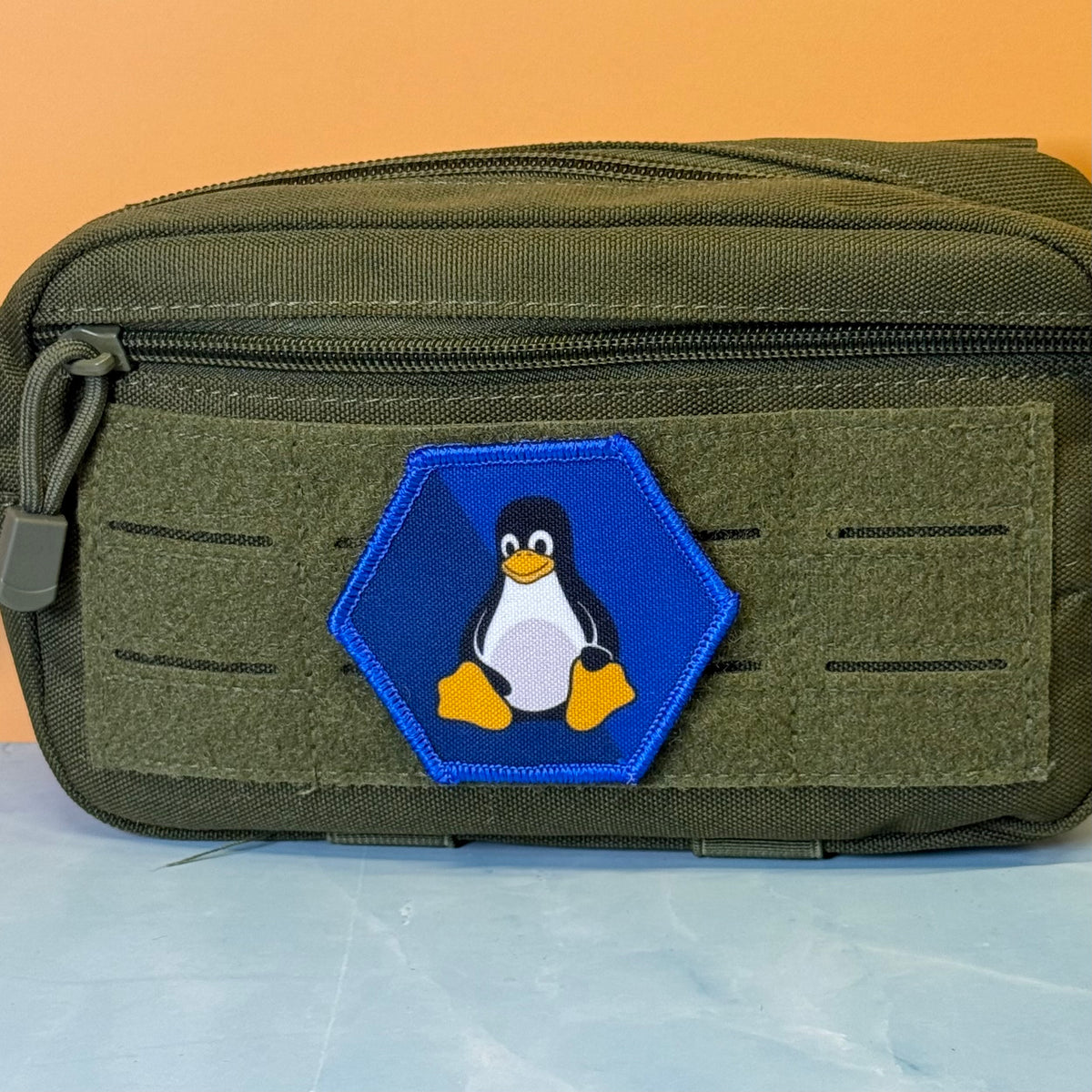Linux Tux Penguin printed canvas morale patch, an olive green tactical pouch with a hexagonal blue patch featuring the Linux Tux penguin logo, set against a split orange and light blue background.