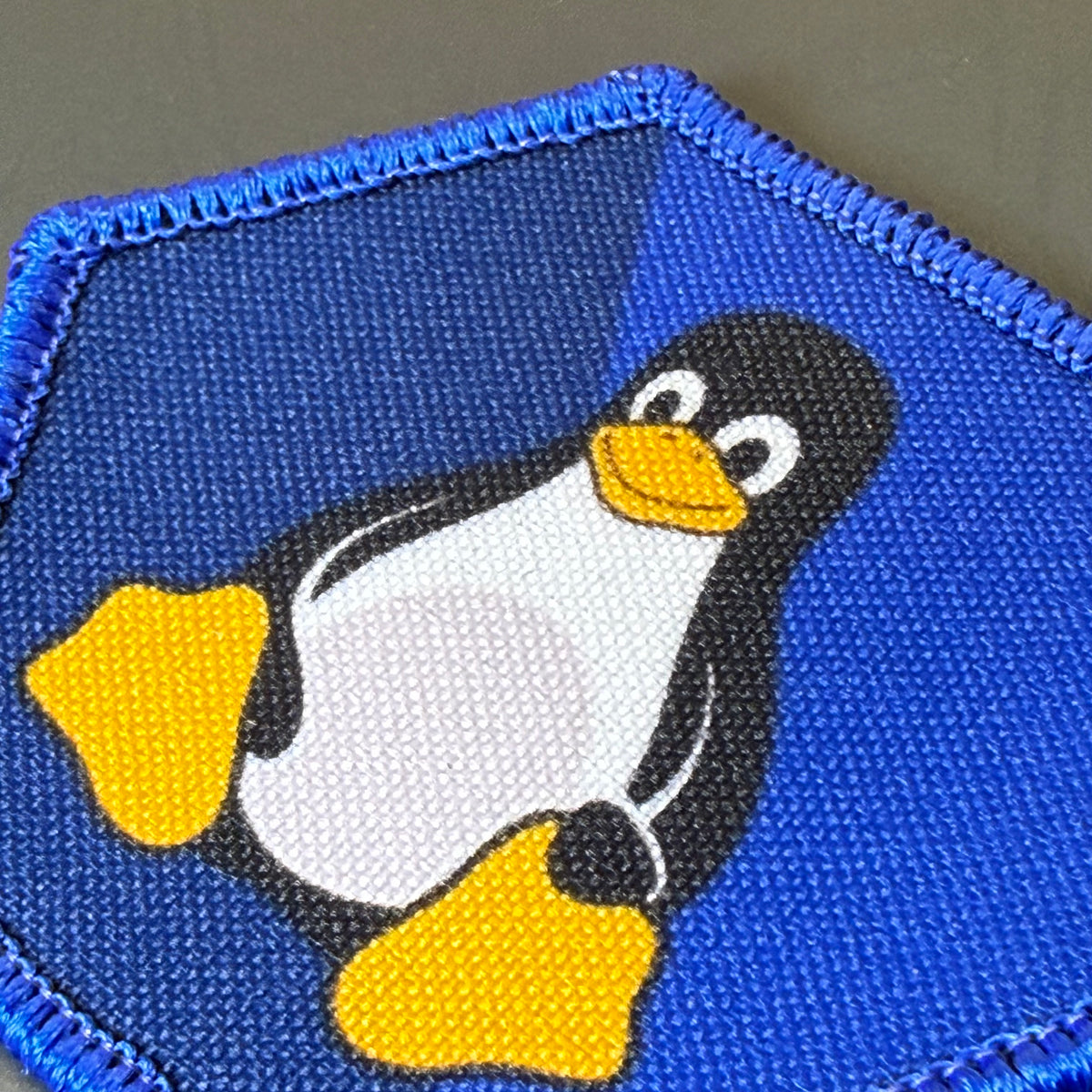 Linux Tux Penguin printed canvas morale patch, a close-up of a blue printed patch featuring the Tuxedo penguin logo with a black body, white belly, and yellow beak and feet on a textured fabric background.