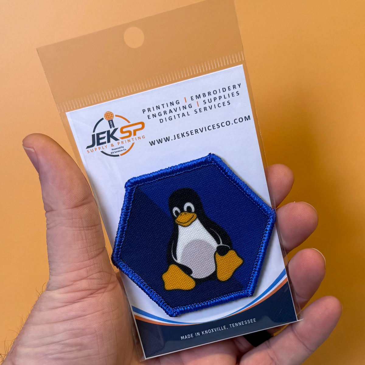 Linux Tux Penguin printed canvas morale patch, a hand holding a clear plastic blister pack containing a hexagonal printed patch with a penguin design. The backing card shows the brand name JEKSP and text for printing.