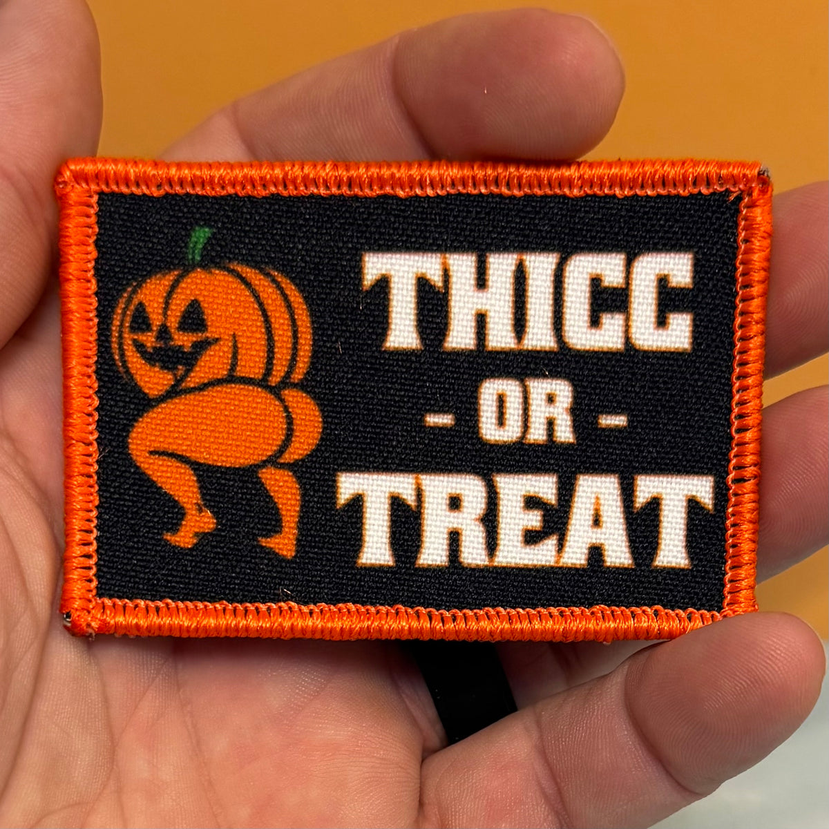 Hand holding the Thicc or Treat printed Halloween pumpkin patch, with cream lettering, a black background and orange border.