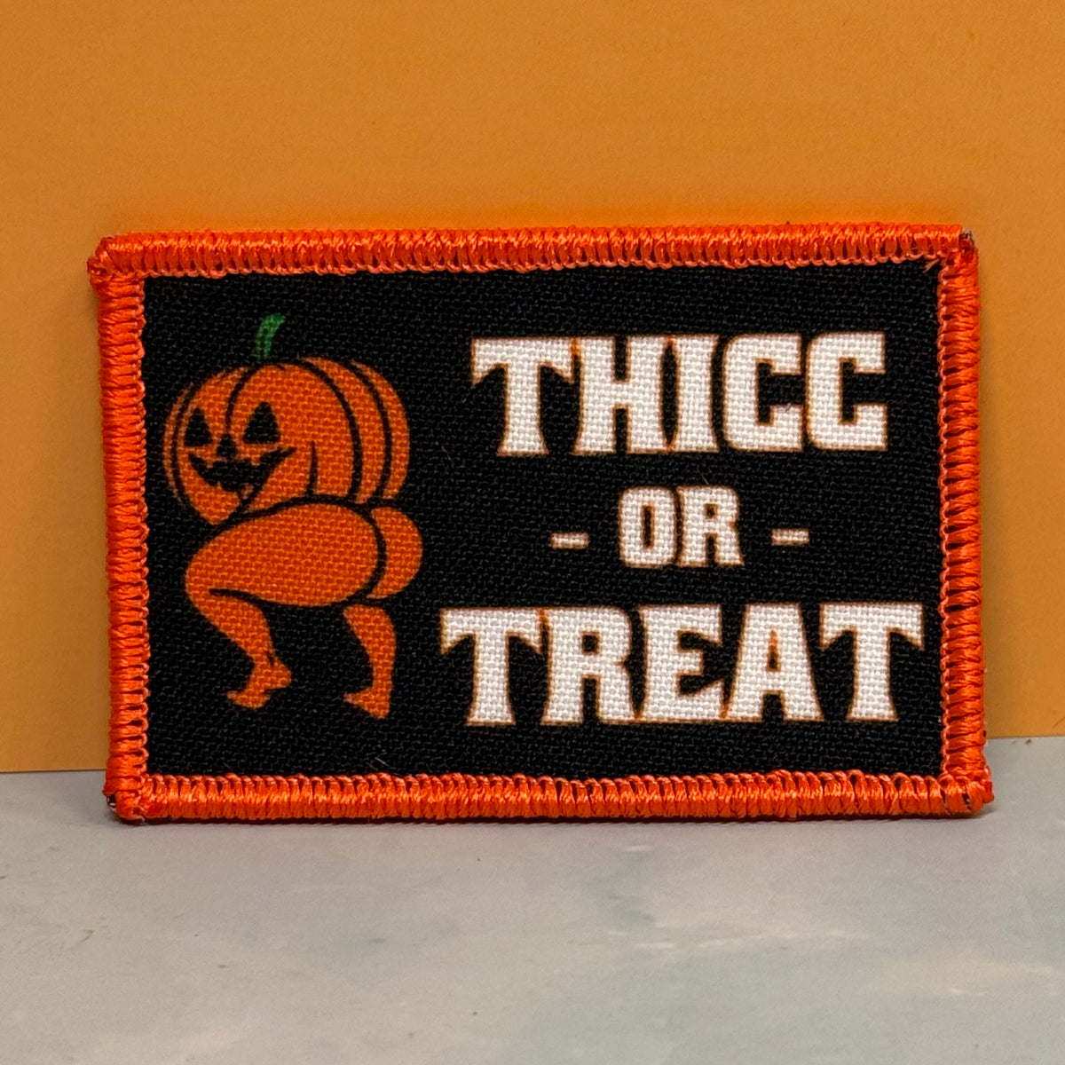 Thicc or Treat printed pumpkin morale patch with orange border photographed against an orange wall and light gray surface.
