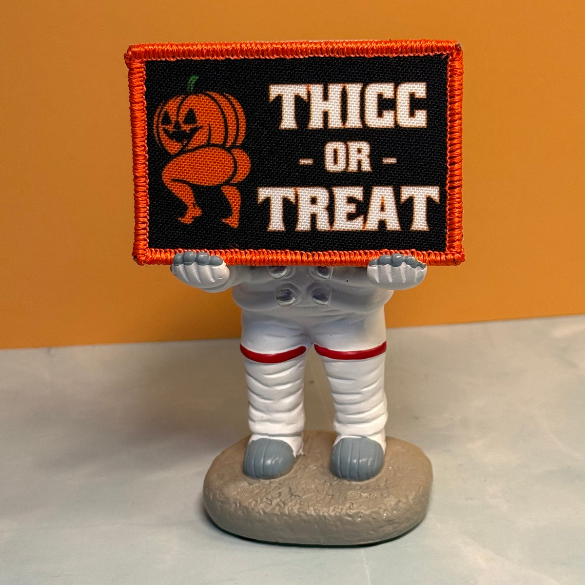 Mummy figurine holding the Thicc or Treat Halloween pumpkin printed morale patch with an orange embroidered border.