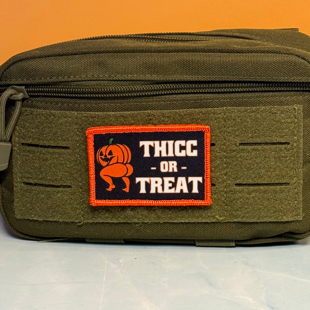 Olive green tactical pouch displaying the Thicc or Treat Halloween pumpkin patch against an orange background.