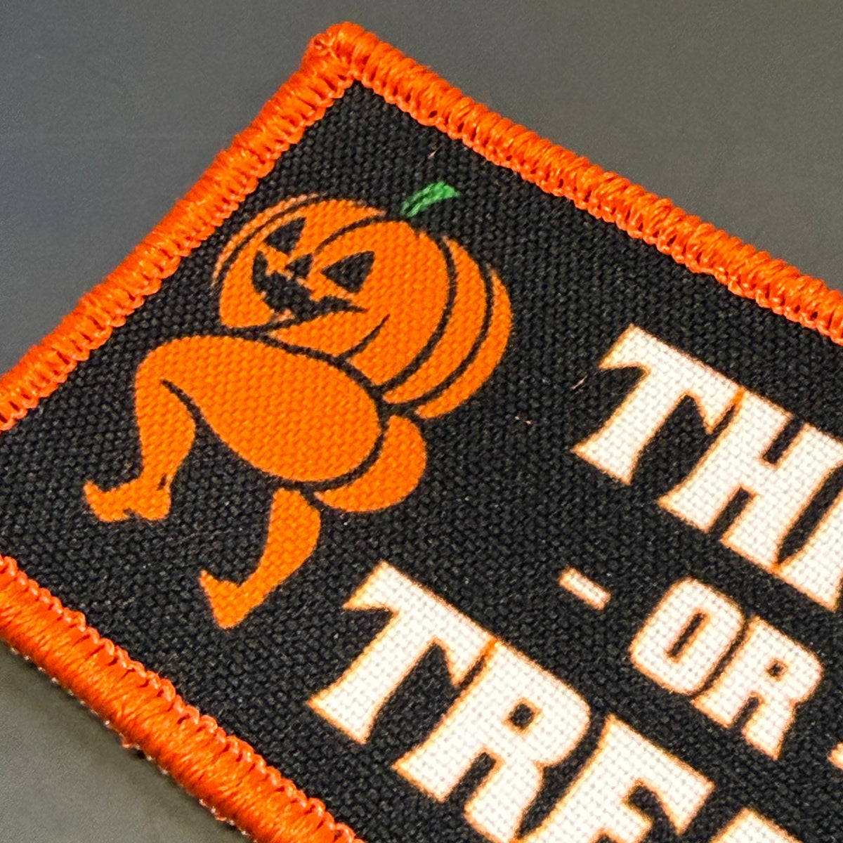 Close-up of the Thicc or Treat printed pumpkin patch showing Halloween artwork, cream lettering and orange embroidered edge.
