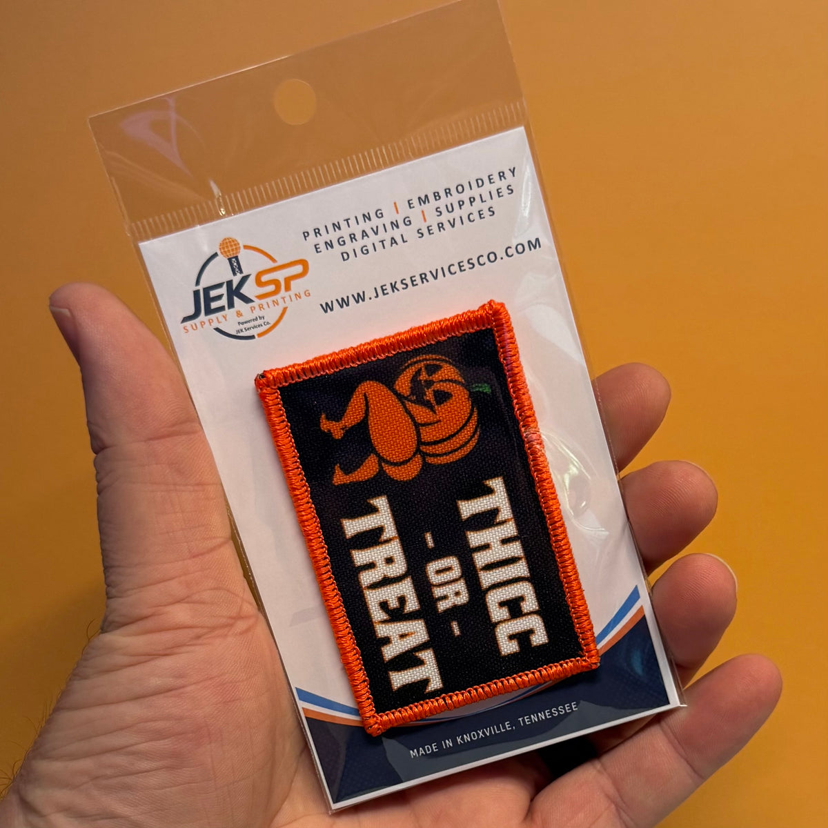 Hand holding a packaged Thicc or Treat Halloween pumpkin printed morale patch with an orange embroidered border.