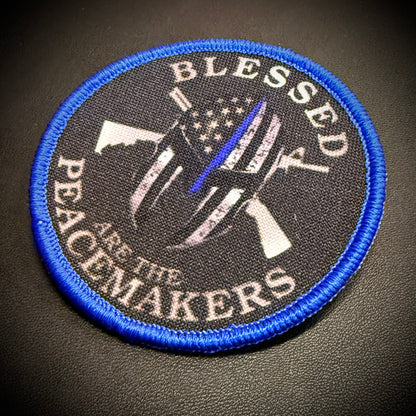 A circular embroidered patch with a blue border and a black background, featuring a stylized peacekeeper helmet with an American flag and rifle, surrounded by the text 'BLESSED PEACEMAKERS'.