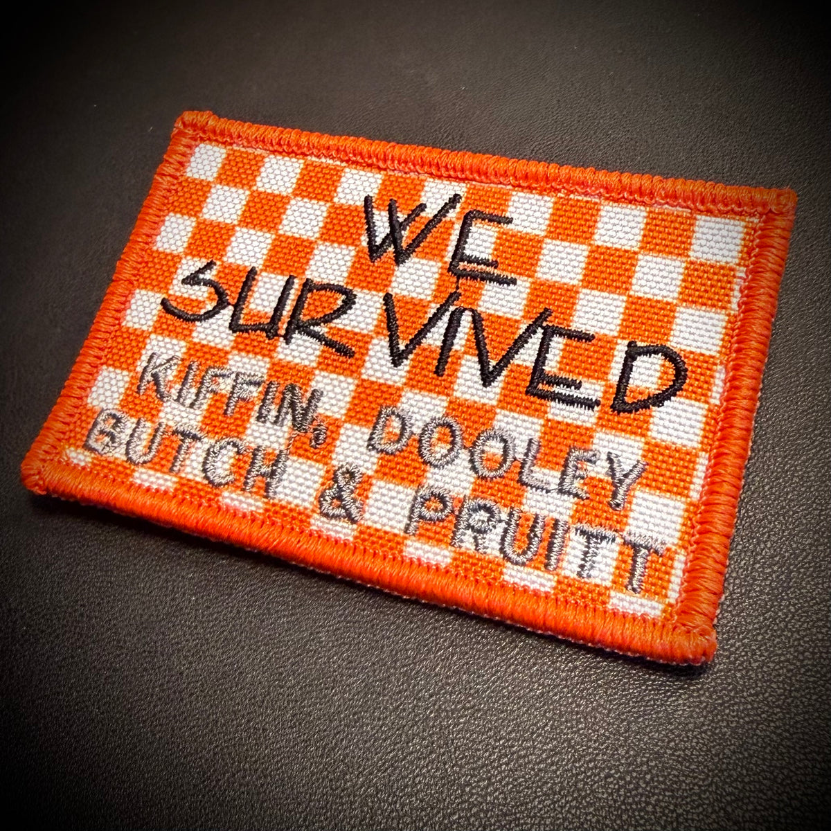 An embroidered rectangular patch with an orange and white checkered border and background, featuring black text that reads 'WE SURVIVED KIFFIN, DOOLEY BUTCH & PRUITT' on a dark textured surface.