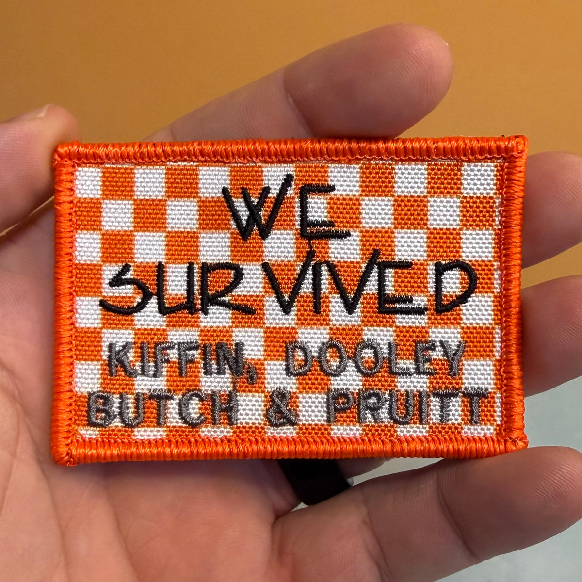 We Survived, Tennessee Football embroidered morale patch: held in a hand, featuring an orange and white checkerboard pattern with black and gray text reading 'WE SURVIVED KIFFIN, DOOLEY BUTCH & PRUITT'.