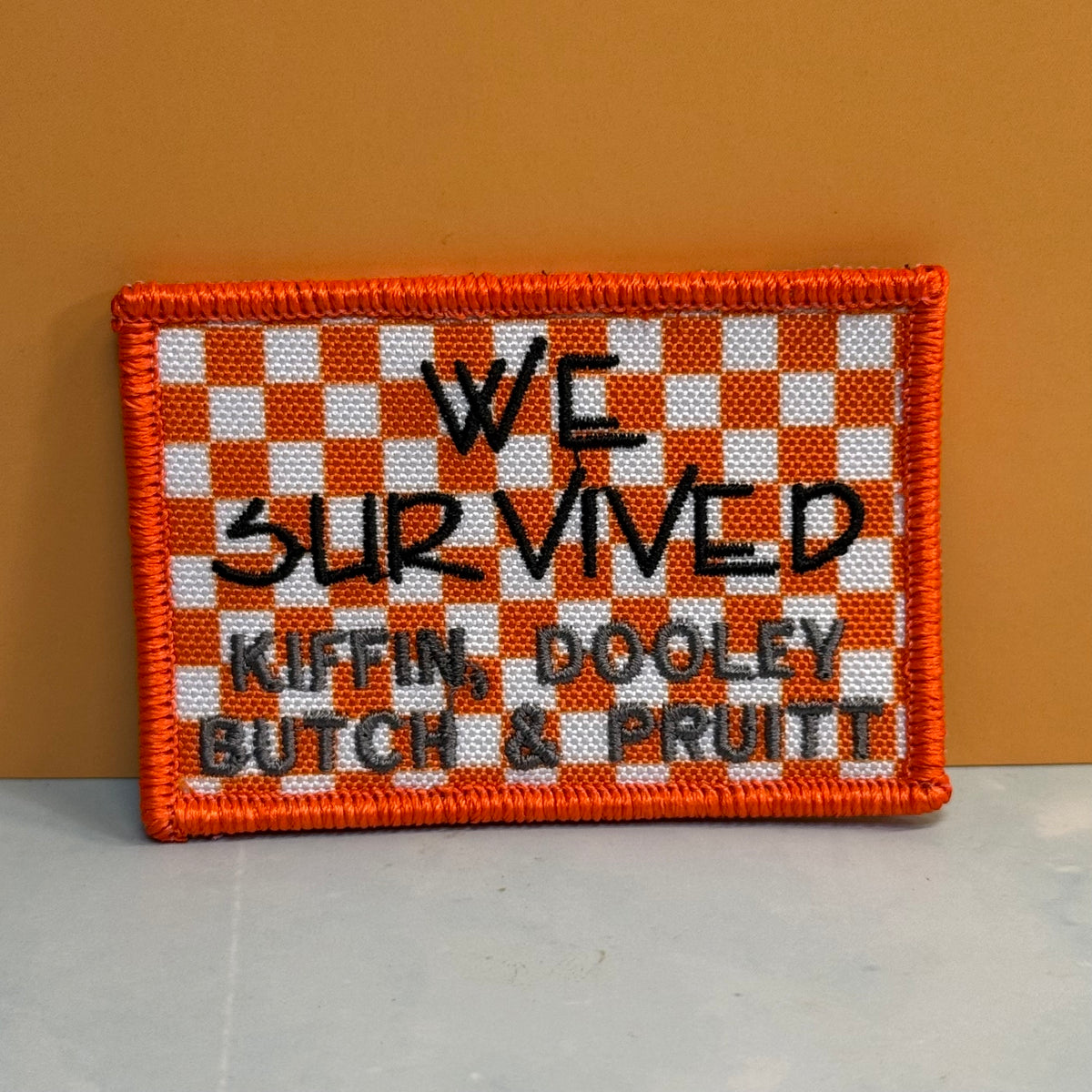 We Survived, Tennessee Football embroidered morale patch: rectangular patch with an orange and white checkerboard pattern and black text reading 'WE SURVIVED KIFFIN, DOOLEY BUTCH & PRUITT', shown against a warm orange-brown.