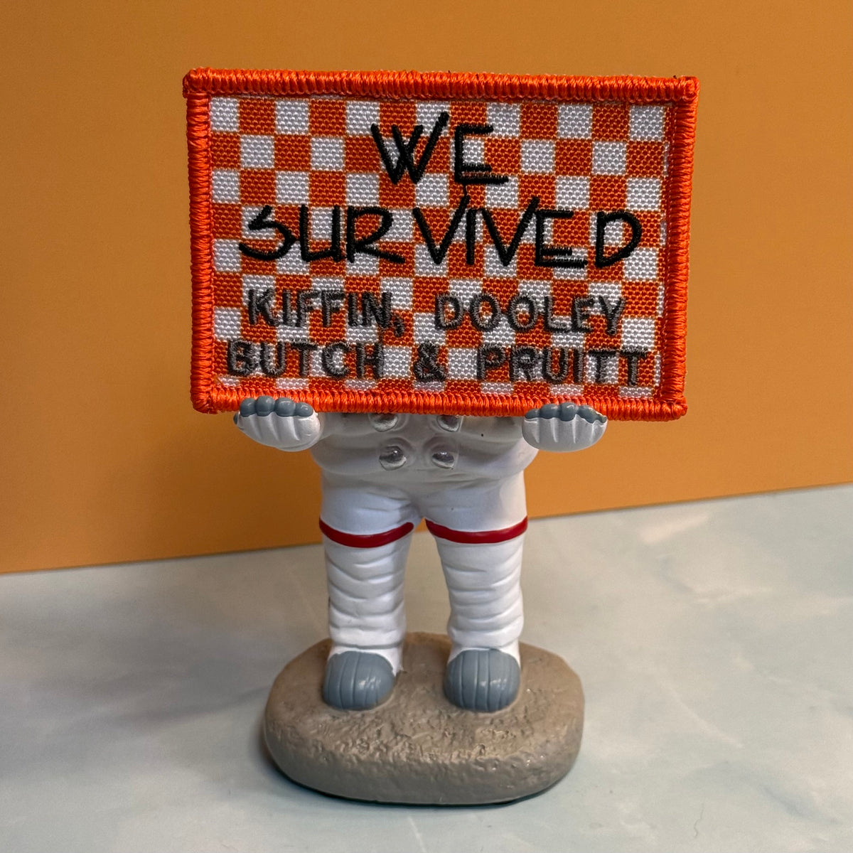 We Survived, Tennessee Football embroidered morale patch: small astronaut figurine holding a square embroidered sign that reads “WE SURVIVED KIFFIN, DOOLEY, BUTCH & PRUITT.” The sign has an orange and white checkered border.