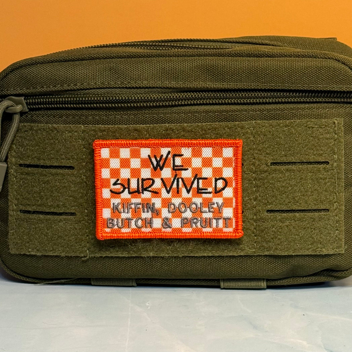 We Survived, Tennessee Football embroidered morale patch: olive green tactical pouch with a rectangular orange and white checkered patch that reads 'WE SURVIVED KIFLIN, DOOLEY, BUTCH & PRUITT'.