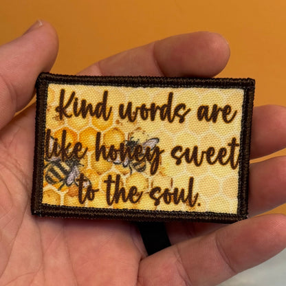 A rectangular embroidered patch held in a hand, featuring the quote 'Kind words are like honey sweet to the soul.' in dark brown script over a yellow honeycomb and bee design, with a dark brown border.