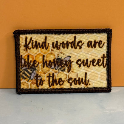 A rectangular embroidered patch with a dark brown border and the phrase 'Kind words are like honey sweet to the soul.' in dark brown script over a honeycomb and bee design, displayed against an orange background.