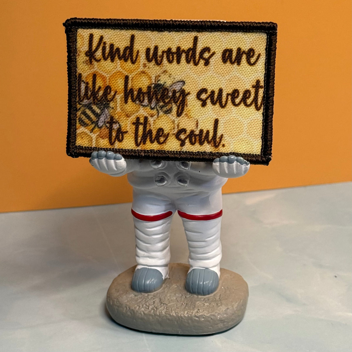 A small figurine of a person in a white astronaut suit holding a rectangular embroidered patch that reads, 'Kind words are like honey sweet to the soul.' The background is a solid orange wall with a light gray surface below.