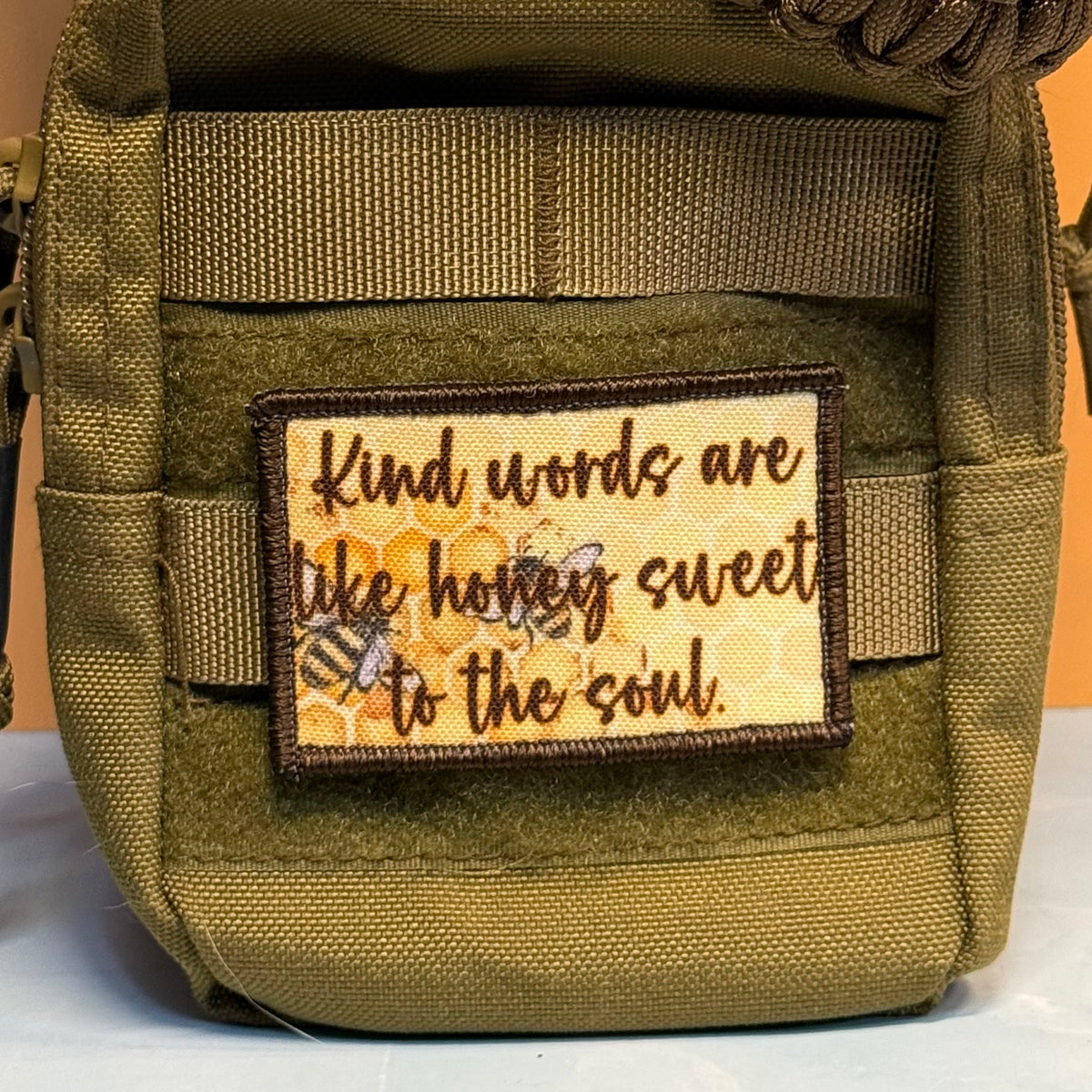 An olive green tactical pouch with a rectangular embroidered patch featuring the quote 'Kind words are like honey sweet to the soul' in brown script over a yellow honeycomb and bee design.