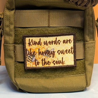 An olive green tactical pouch with a rectangular embroidered patch featuring the quote 'Kind words are like honey sweet to the soul' in brown script over a yellow honeycomb and bee design.