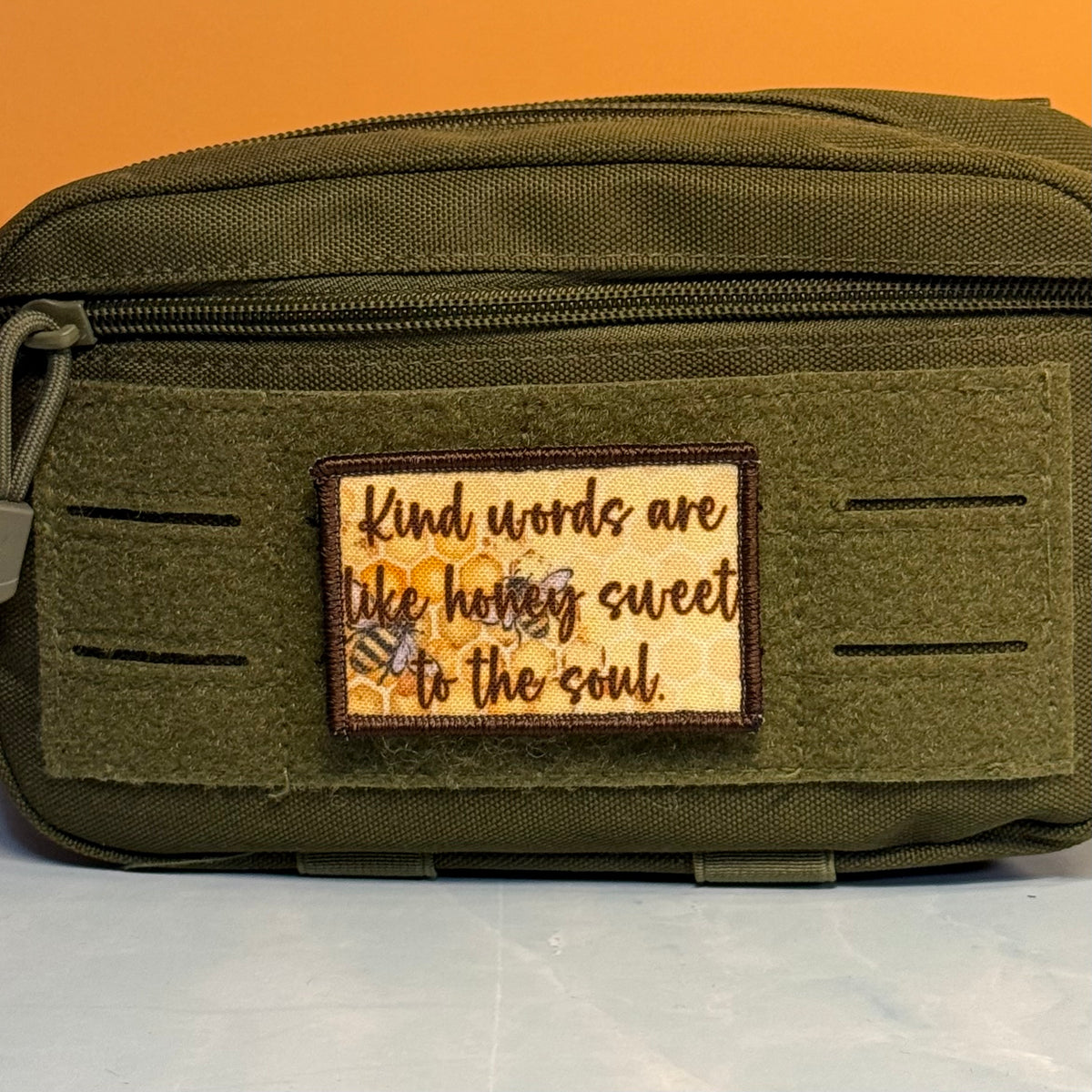 An olive green tactical pouch with a front hook-and-loop patch that reads 'Kind words are like honey sweet to the soul.' with bees and honeycomb details, set against an orange background.