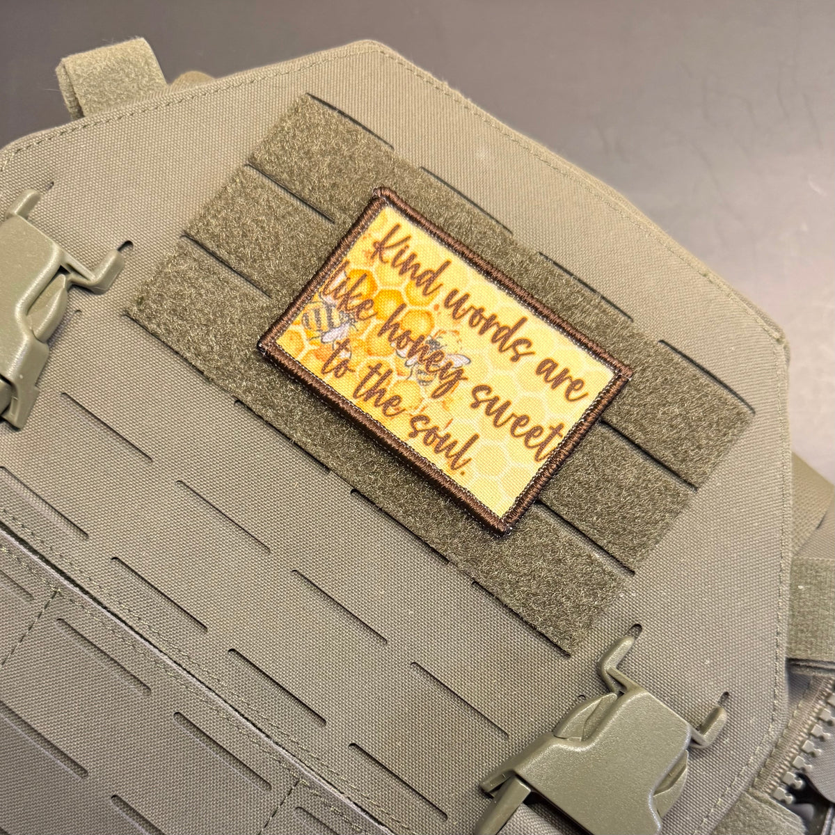 A rectangular embroidered patch with a honeybee and the quote 'Kind words are the honey sweet to the soul.' in brown lettering on a yellow honeycomb background, attached to olive drab tactical webbing on a bag.