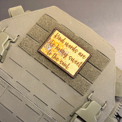 A rectangular embroidered patch with a honeybee and the quote 'Kind words are the honey sweet to the soul.' in brown lettering on a yellow honeycomb background, attached to olive drab tactical webbing on a bag.