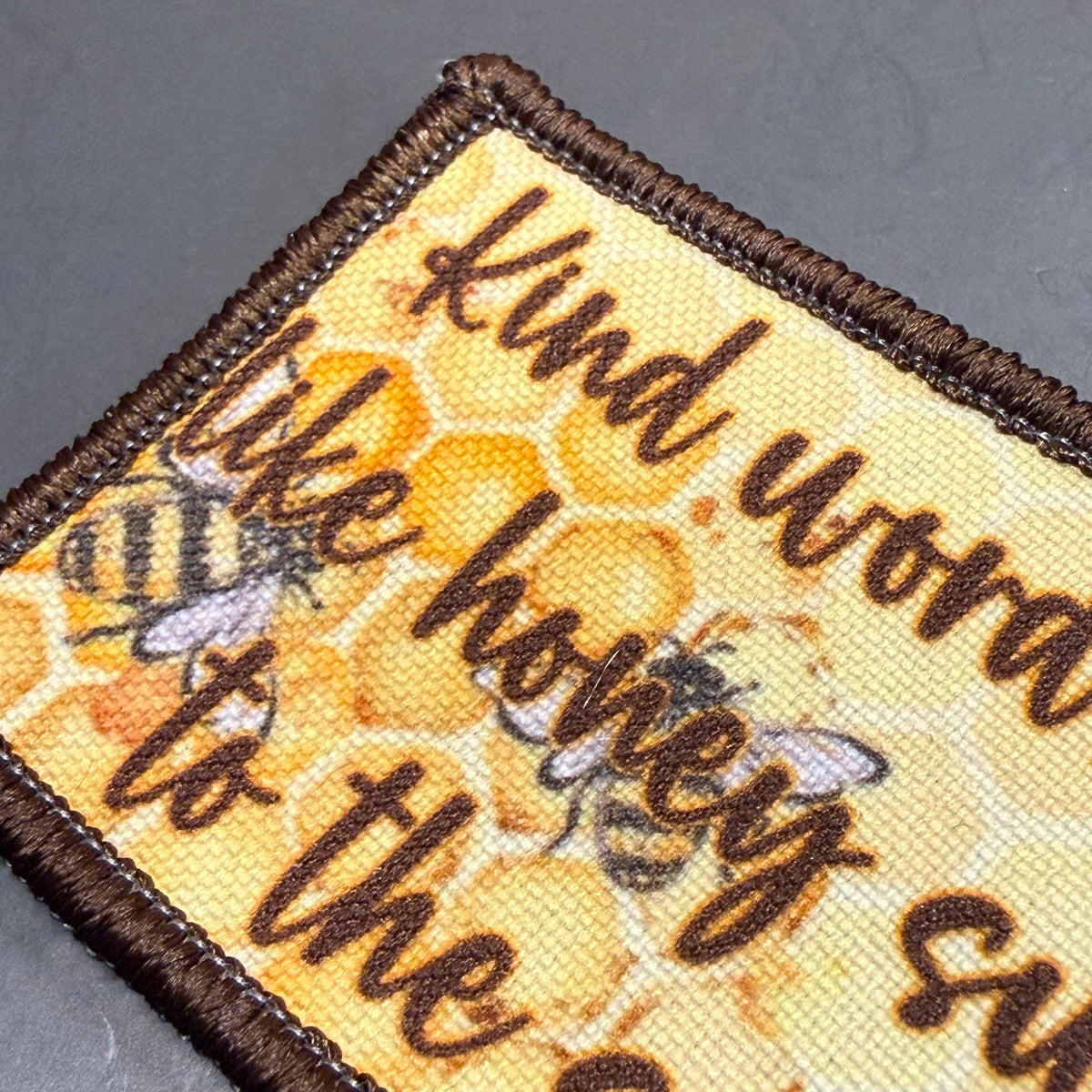 A close-up of a rectangular embroidered patch with a brown stitched border, featuring a honeycomb pattern, bees, and the phrase 'Kind words like honey suits the bee' in dark brown script on a yellow background.