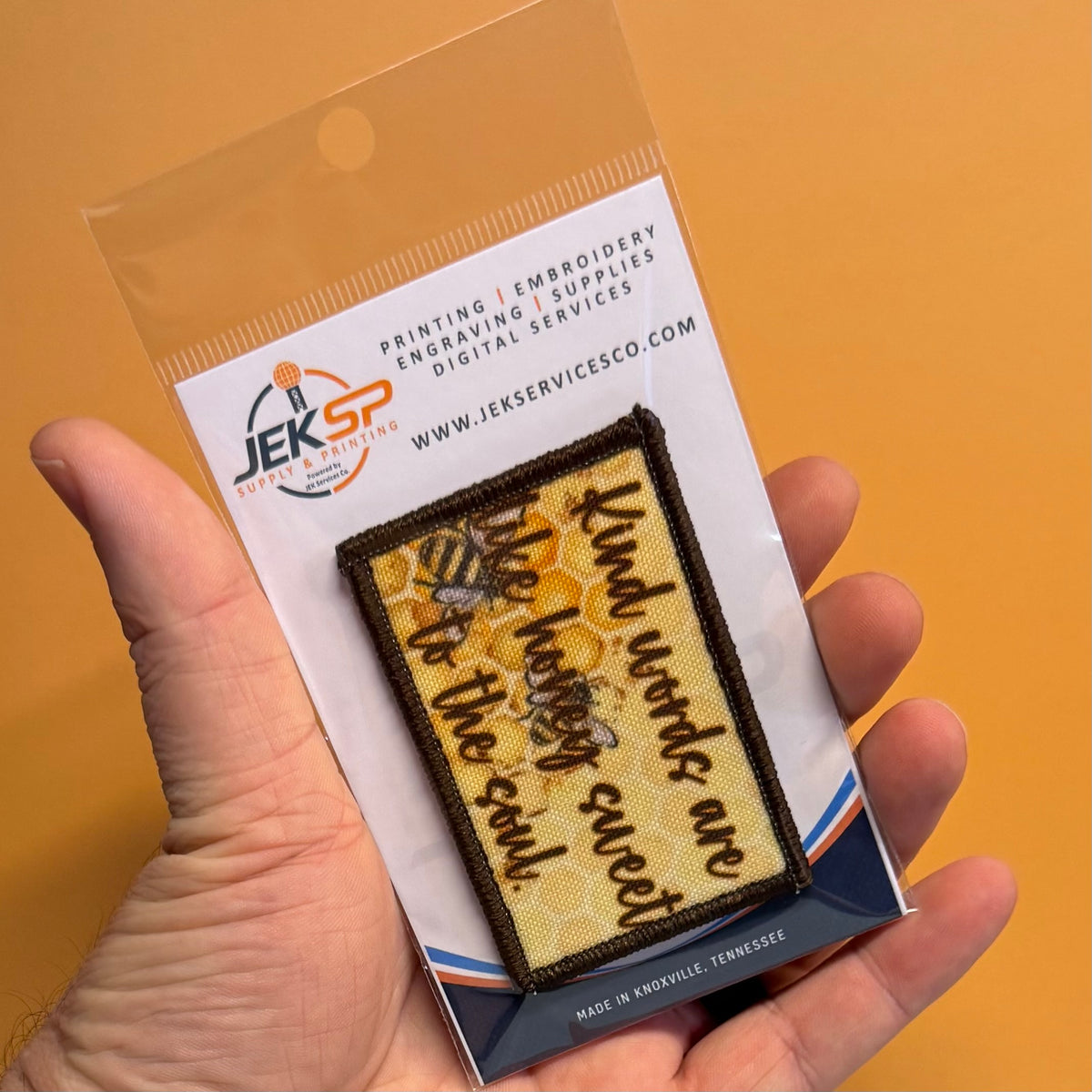 A hand holding a clear plastic package containing a rectangular embroidered patch. The patch has a yellow honeycomb pattern with brown text reading 'kind words are like honey sweet to the soul' and a bee design.