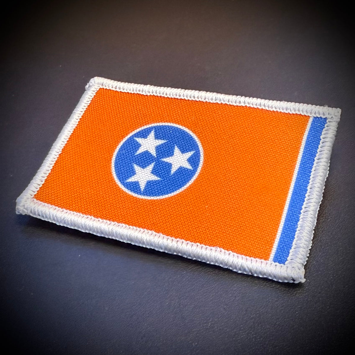 An embroidered rectangular patch of the Tennessee state flag, featuring an orange field, a blue circle with three white stars, and a blue vertical stripe, finished with a white stitched border, shown on a dark textured background.